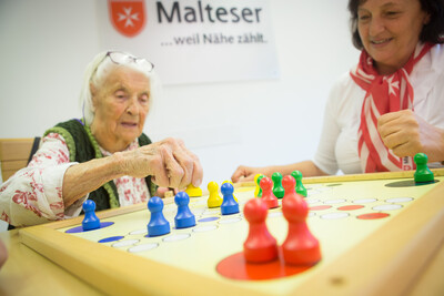 Eine alte Dame spielt mit einer weiteren Frau Mensch ärgere dich nicht. Das Spielbrett und die Spielfiguren sind dabei besonders groß gestaltet. 