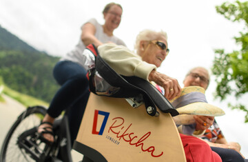 In einer Rikscha sitzen vorne zwei Seniorinnen, das Fahrrad selbst wird von einer Frau gefahren. Miteinander genießen sie das gemächliche Tempo, den Ausblick und die Gespräche.