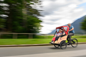 Eine Rikscha fährt mit zwei Seniorinnen und einem Fahrer eine Straße entlang. Im Hintergrund sind Wolken, Berge, Bäume und Wiese zu sehen. 
