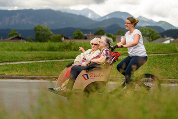 Drei Frauen fahren mit einer E-Rikscha. Die beiden Seniorinnen sitzen vorne und unterhalten sich mit der jungen Frau, die die Rikscha fährt. Im Hintergrund sind die Berge zu sehen, davor grüne Wiesen und Hausdächer.